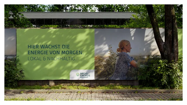 Ein großes Werbeplakat an einem Zaun im Freien mit der Aufschrift „HIER WÄCHST DIE ENERGIE VON MORGEN. LOKAL & NACHHALTIG.“ der Marke Regio Energie Musterort.