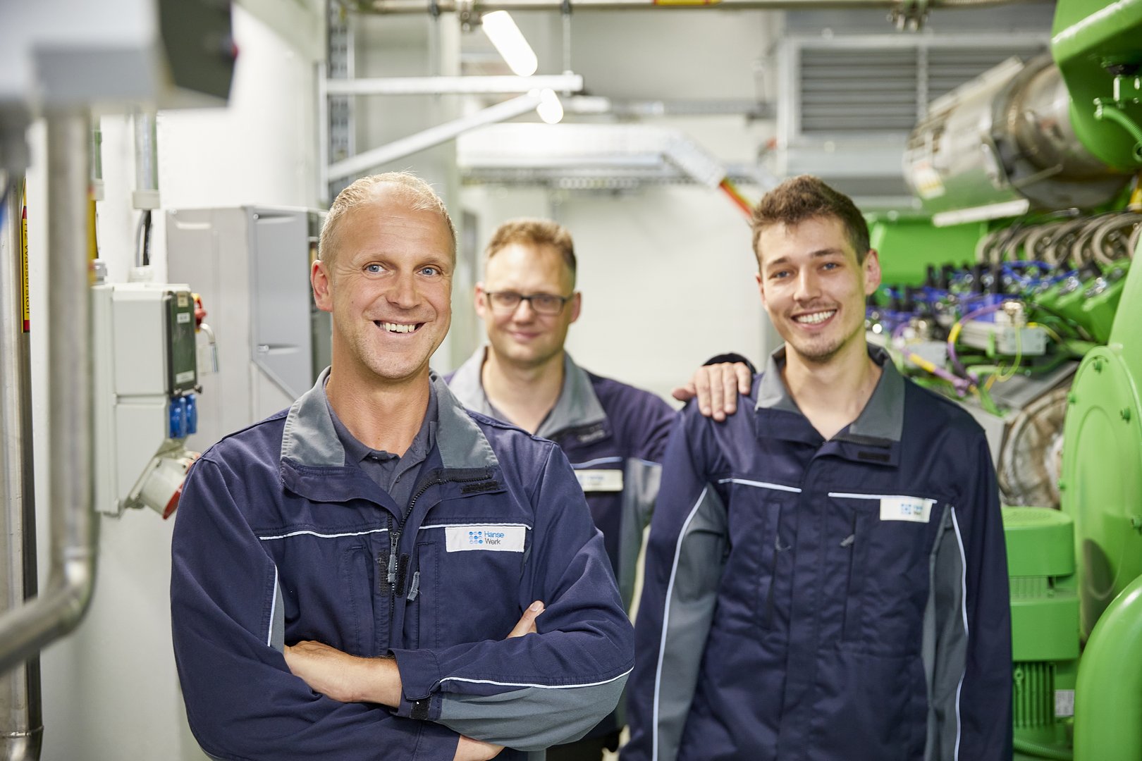 Drei lächelnde Techniker in Arbeitskleidung der HanseWerk-Gruppe in einem Technikraum für Energieversorgung.