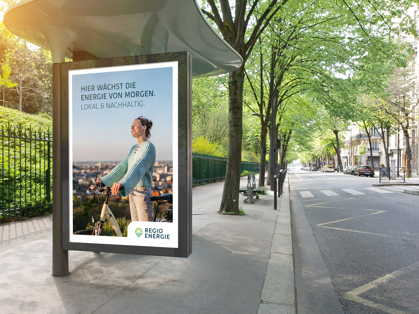 Ein digitales Werbeplakat von RegioEnergie mit einer Frau und Fahrrad steht an einem Baum-gesäumten Gehweg in einer Stadt.